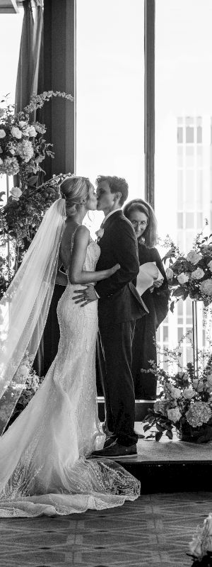 A wedding party gathers in a sunlit room with guests seated, a bride and groom at center, and musicians or attendants nearby, celebrating.