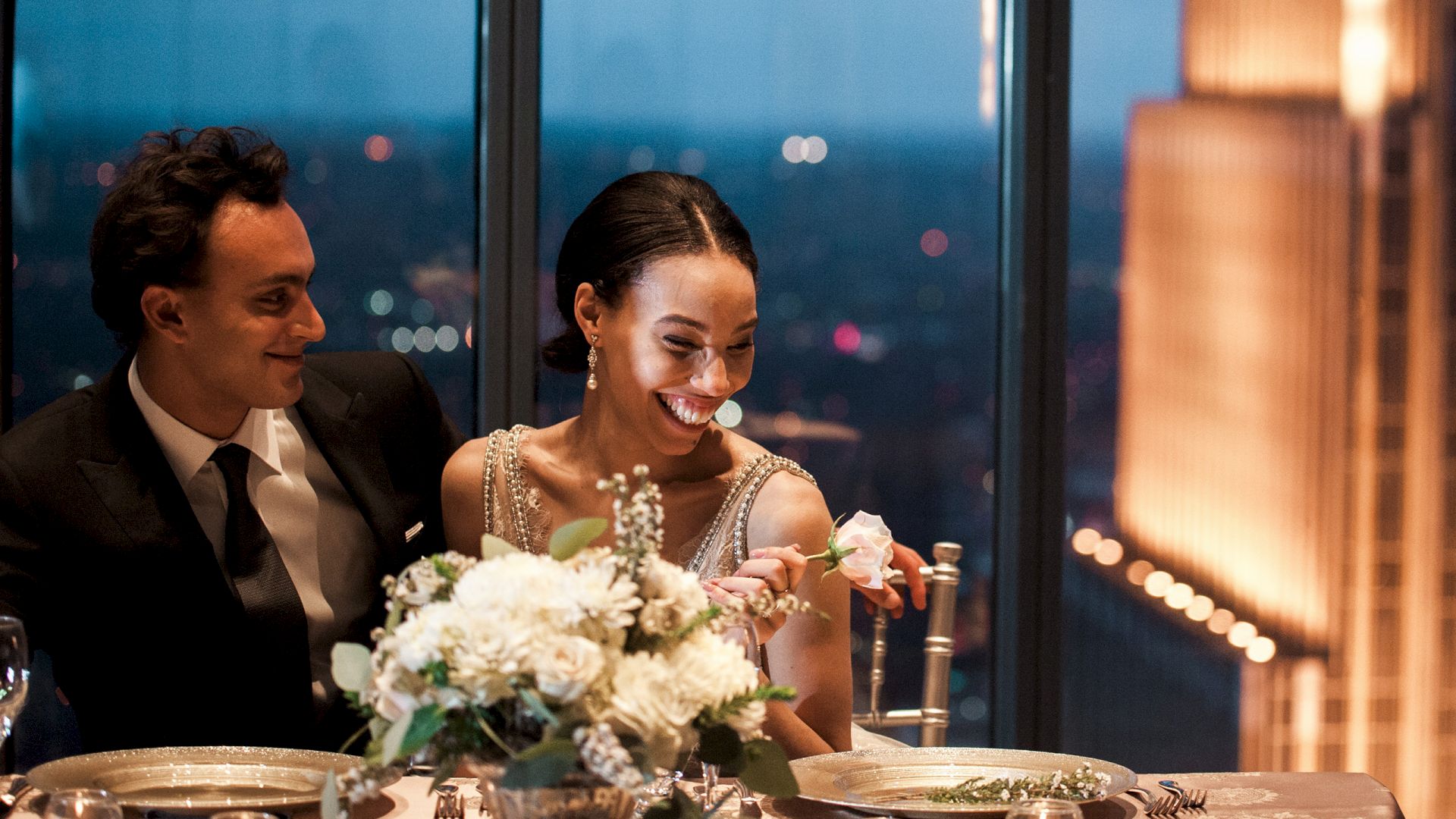 A smiling bride and groom sit at a decorated banquet table, toasting while flowers glow by the window.