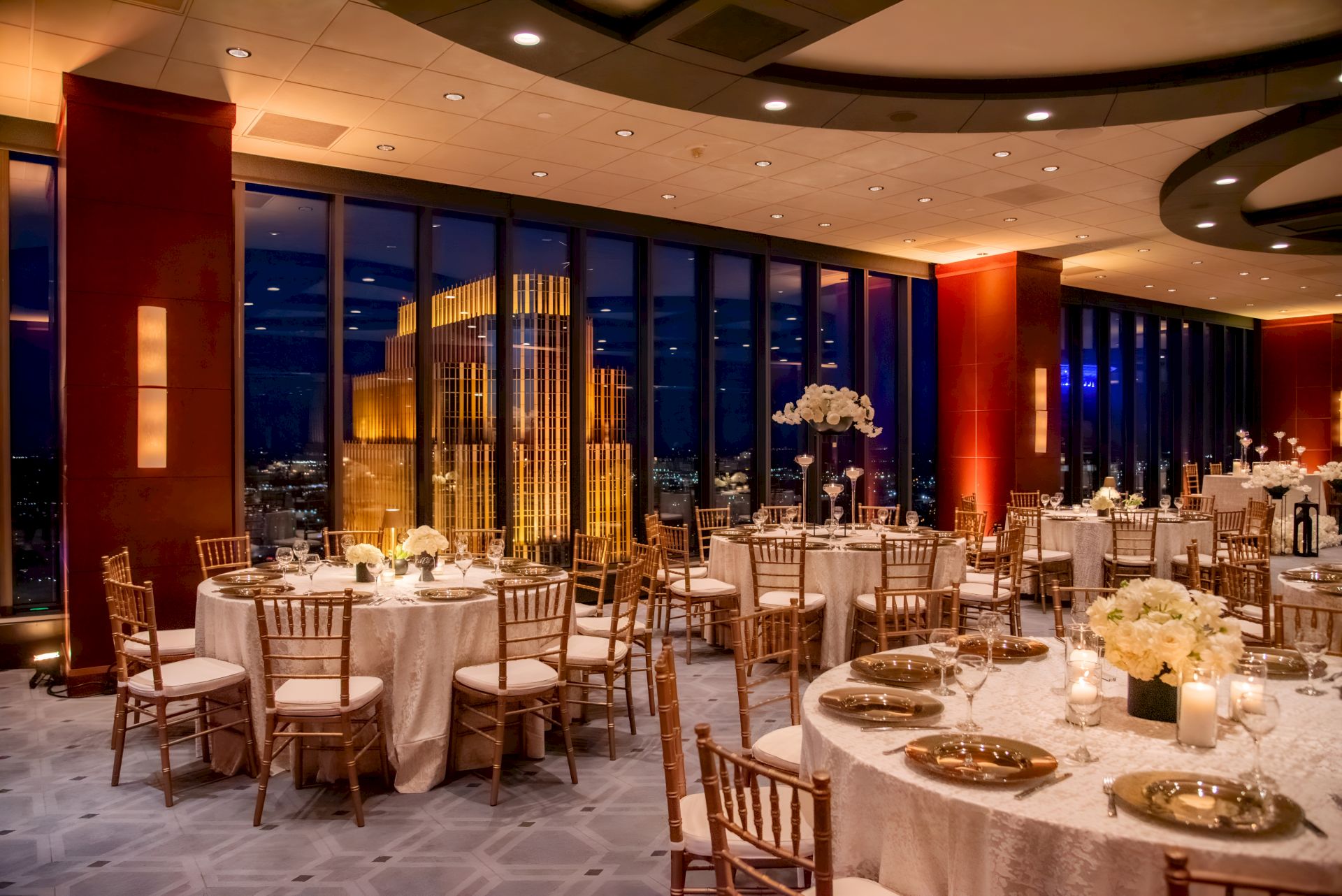 An elegant banquet setup with round tables, gold chairs, candles and flowers, overlooking a night city through tall windows under warm lights.