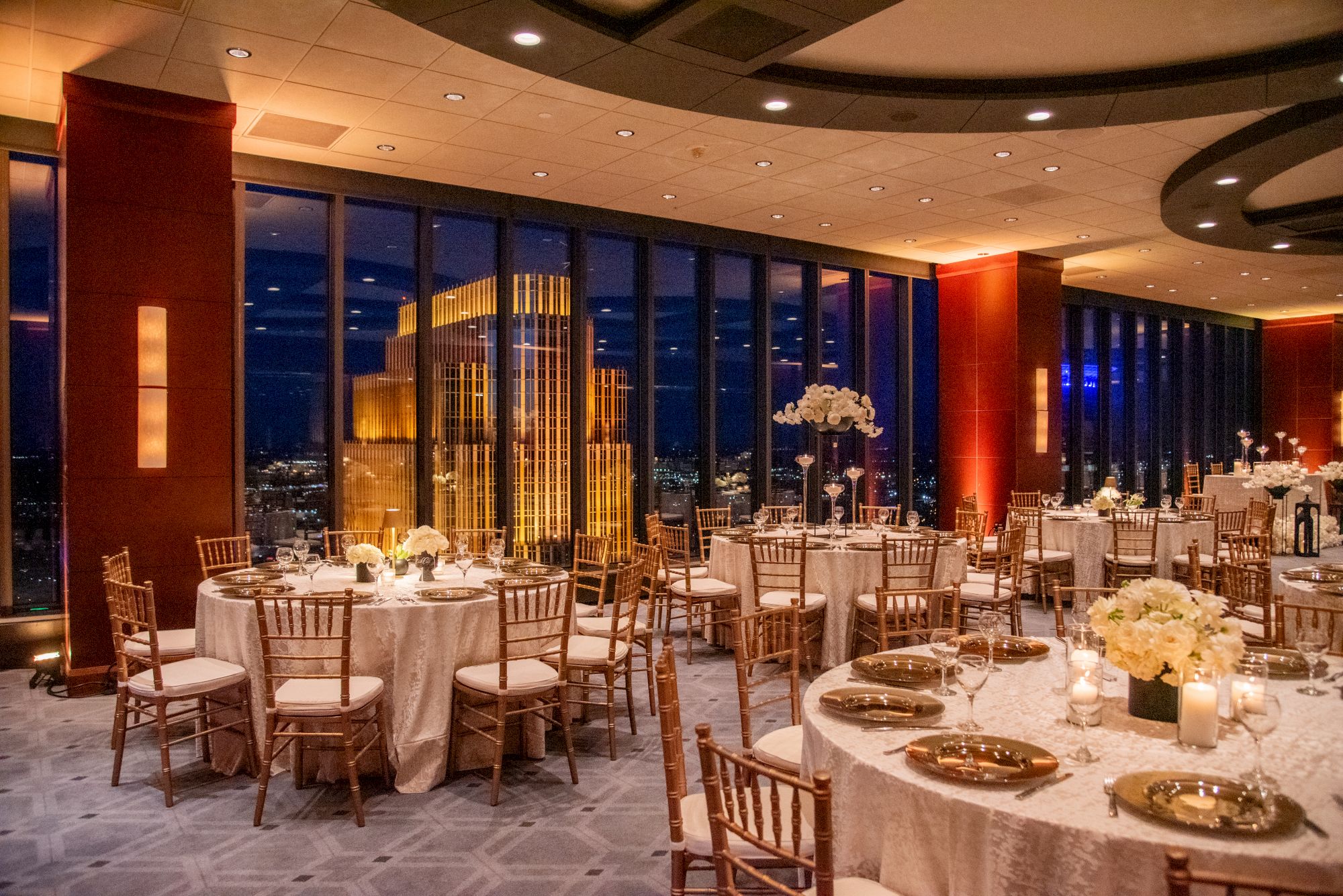 A formal banquet hall with round tables, white linens, gold chairs, candles, floral centerpieces, and large windows showing city lights at night.