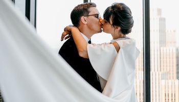 A couple shares a kiss near a window, embraced passionately, with city buildings in the background.