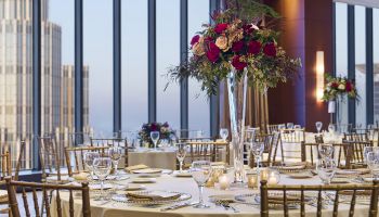 Elegant banquet setup in a high-rise venue: round tables, white linens, crystal glassware, gold chairs, and tall floral centerpieces, city views.