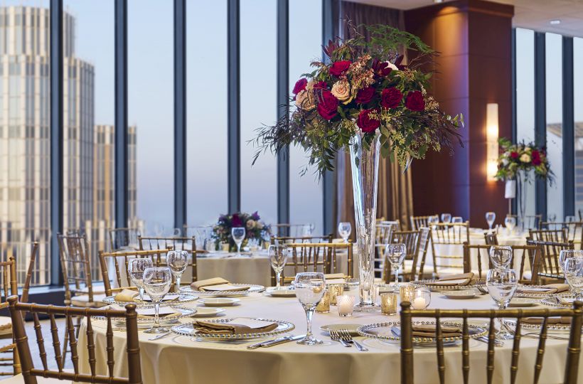Elegant banquet setup in a high-rise venue: round tables, white linens, crystal glassware, gold chairs, and tall floral centerpieces, city views.