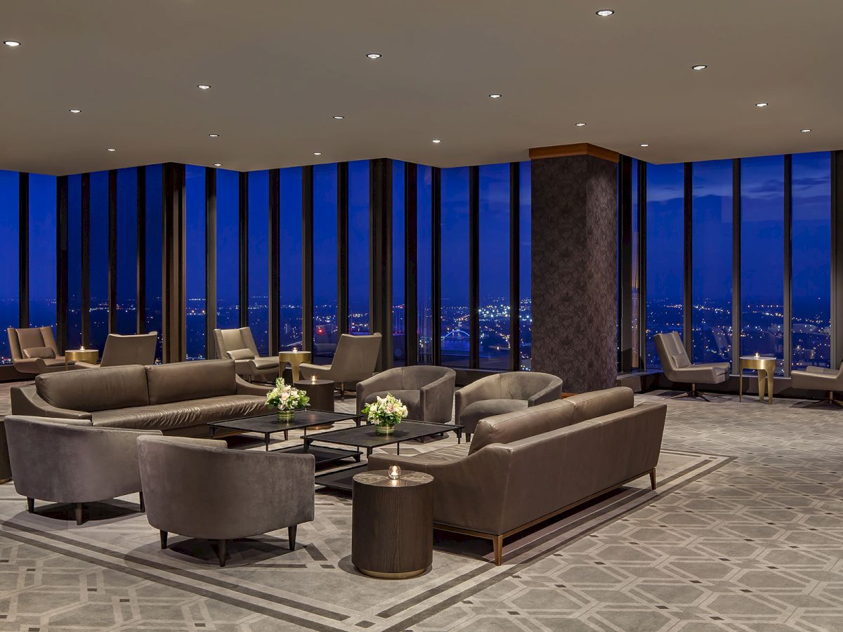A modern, luxurious lounge with gray sofas, round coffee tables, tall windows, night city views, and soft ambient lighting. Ends with a period.
