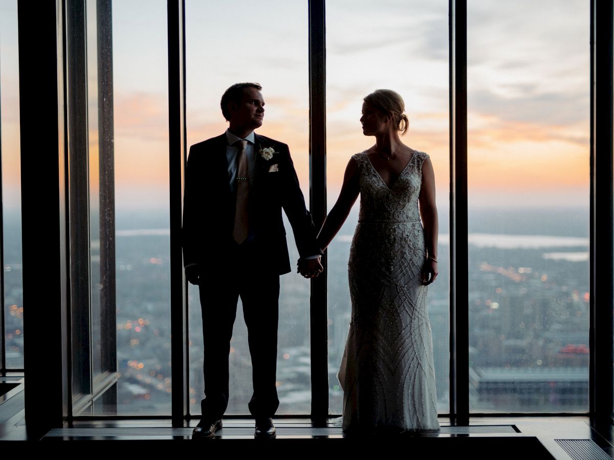A couple in formal attire holds hands by large windows at sunset, city skyline in the background, romantic and elegant moment.