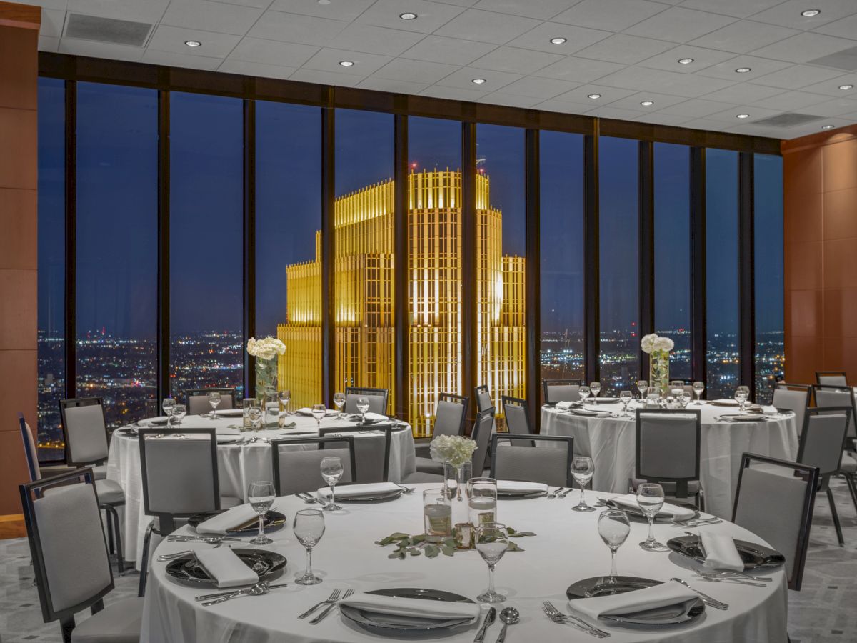 A fancy rooftop restaurant with round tables set for dining, elegant place settings, soft lighting, and a cityscape view at night.