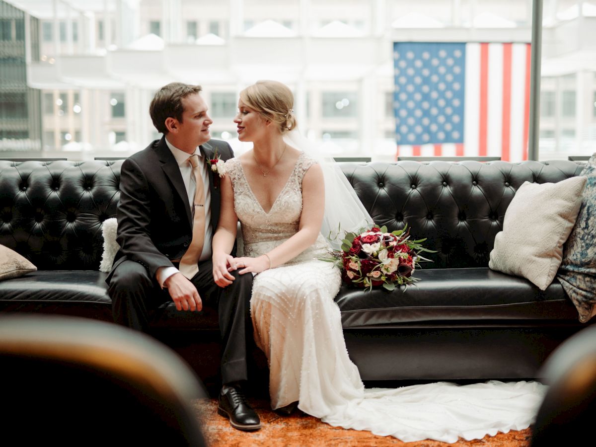 A newlywed couple sits close on a black leather couch, the bride in a white gown holding a bouquet, with a US flag backdrop behind them.