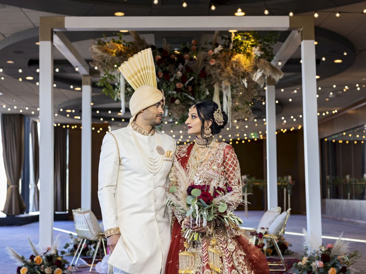 Two people in ornate wedding attire stand under a decorated arch, gazing at each other; the groom wears a tall headpiece, the bride holds flowers.