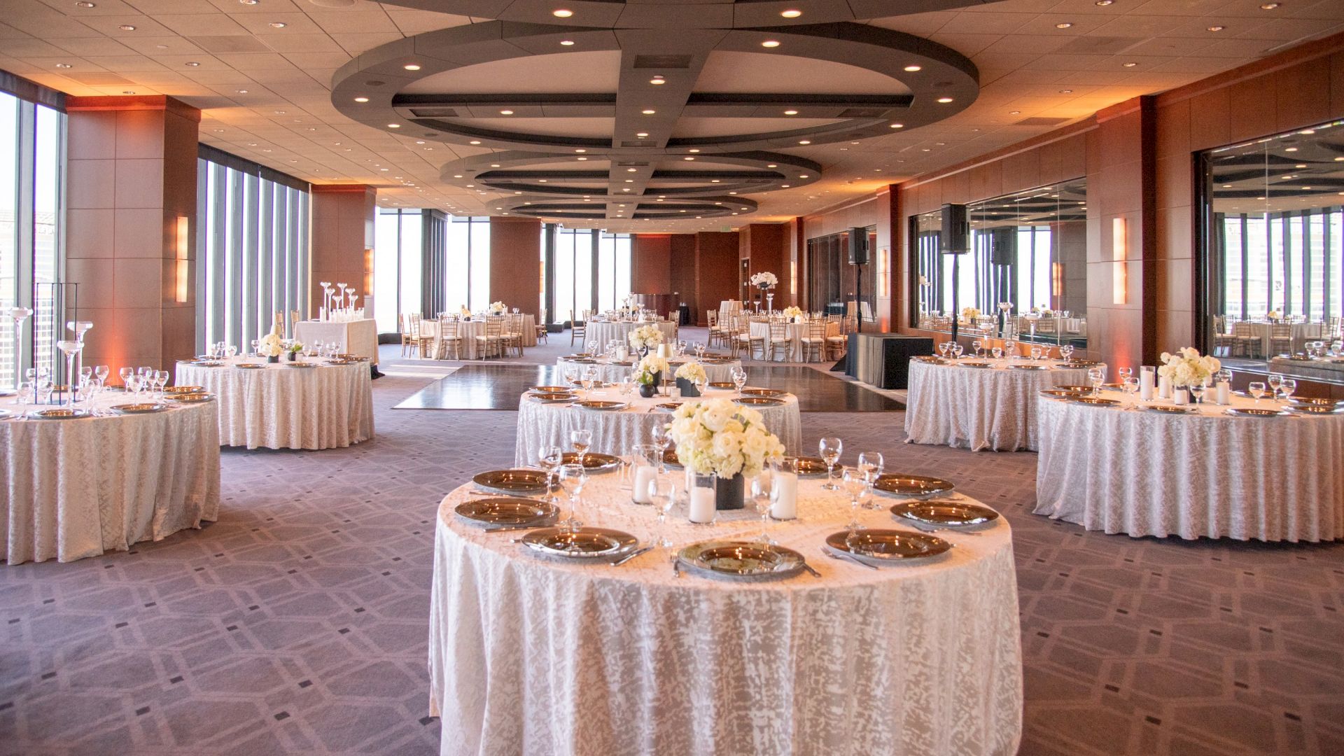 A banquet hall set for a formal event, round tables with white linens, centerpieces, place settings, and soft lighting along windows.