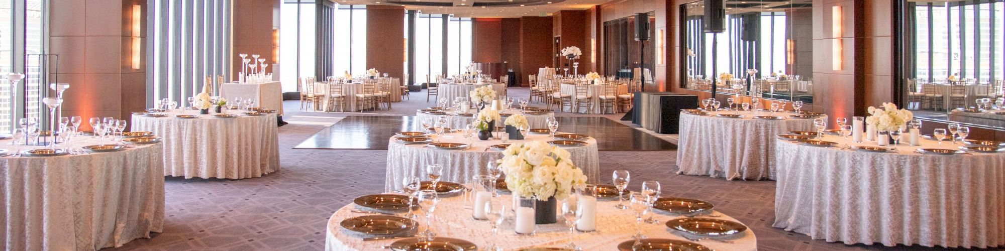 A banquet hall set for a formal event, round tables with white linens, centerpieces, place settings, and soft lighting along windows.