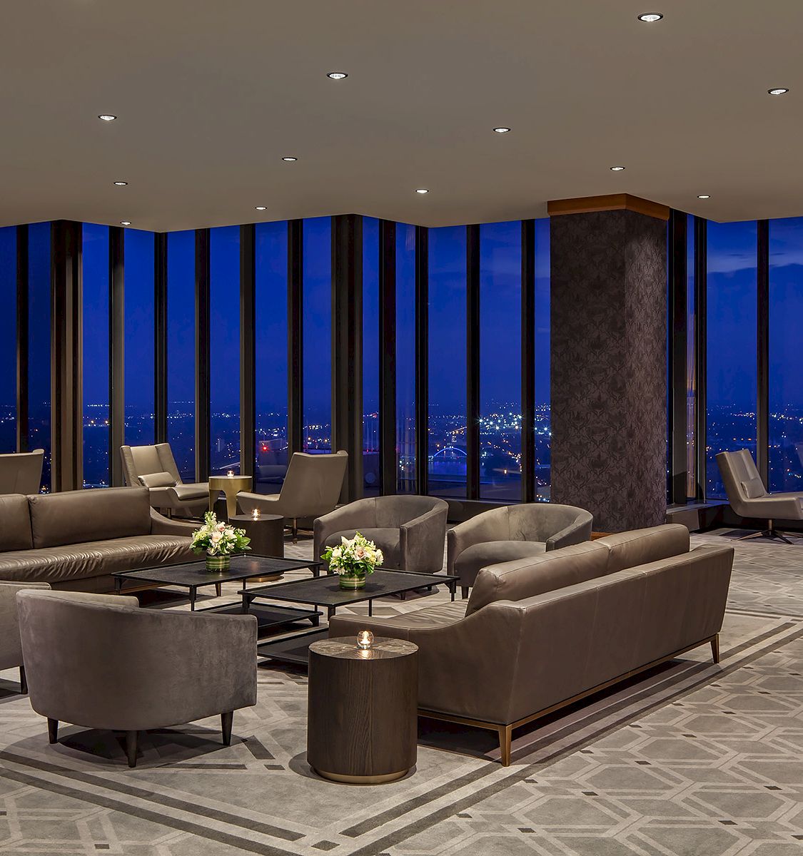A luxurious hotel lounge with plush gray sofas, round tables, and floor-to-ceiling windows offering a night city view, elegant lighting, and calm ambiance.