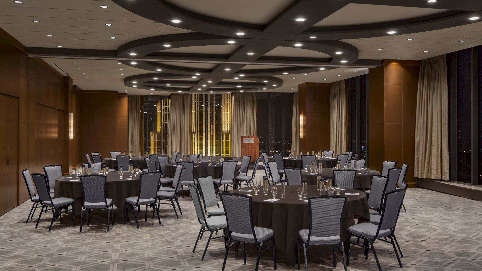 A modern banquet room set with round tables, chairs, and glassware, lit by recessed ceiling lights.