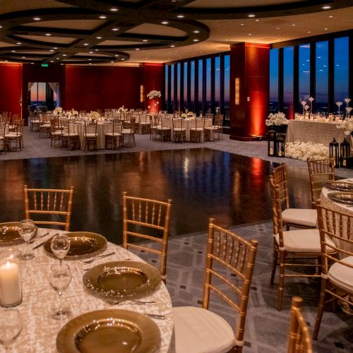 Elegant banquet hall with round tables, gold chairs, white linens, candle decor, and soft warm lighting for a formal event, evening skyline in the background.