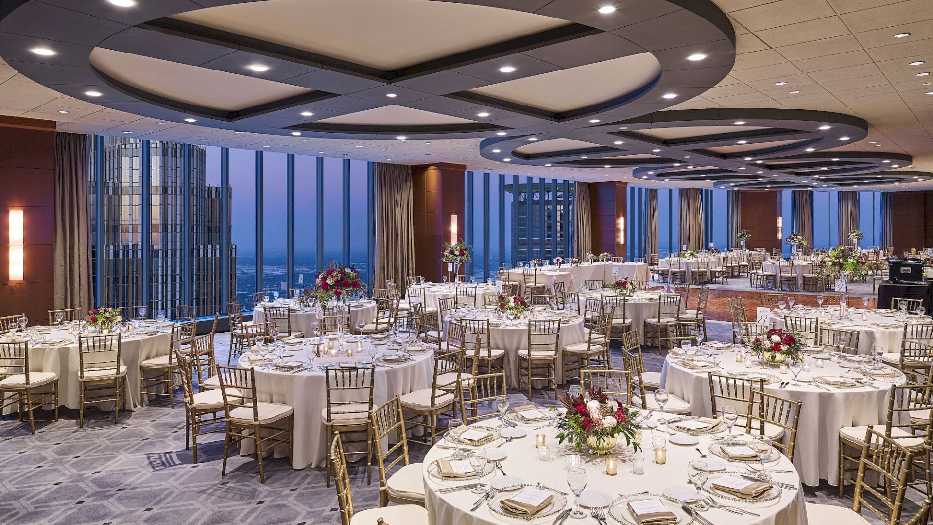 A banquet hall set for a wedding or event, with round tables, floral centerpieces, and gold chairs under modern ring lights.