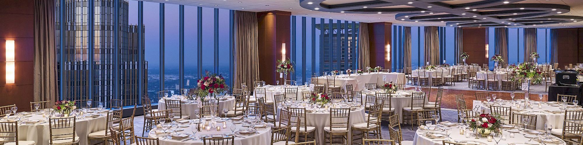 A banquet hall set for a wedding or event, with round tables, floral centerpieces, and gold chairs under modern ring lights.