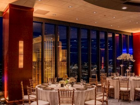 A banquet hall setup with round tables, white tablecloths, gold chairs, elegant place settings, and a cityscape view through large windows at night.