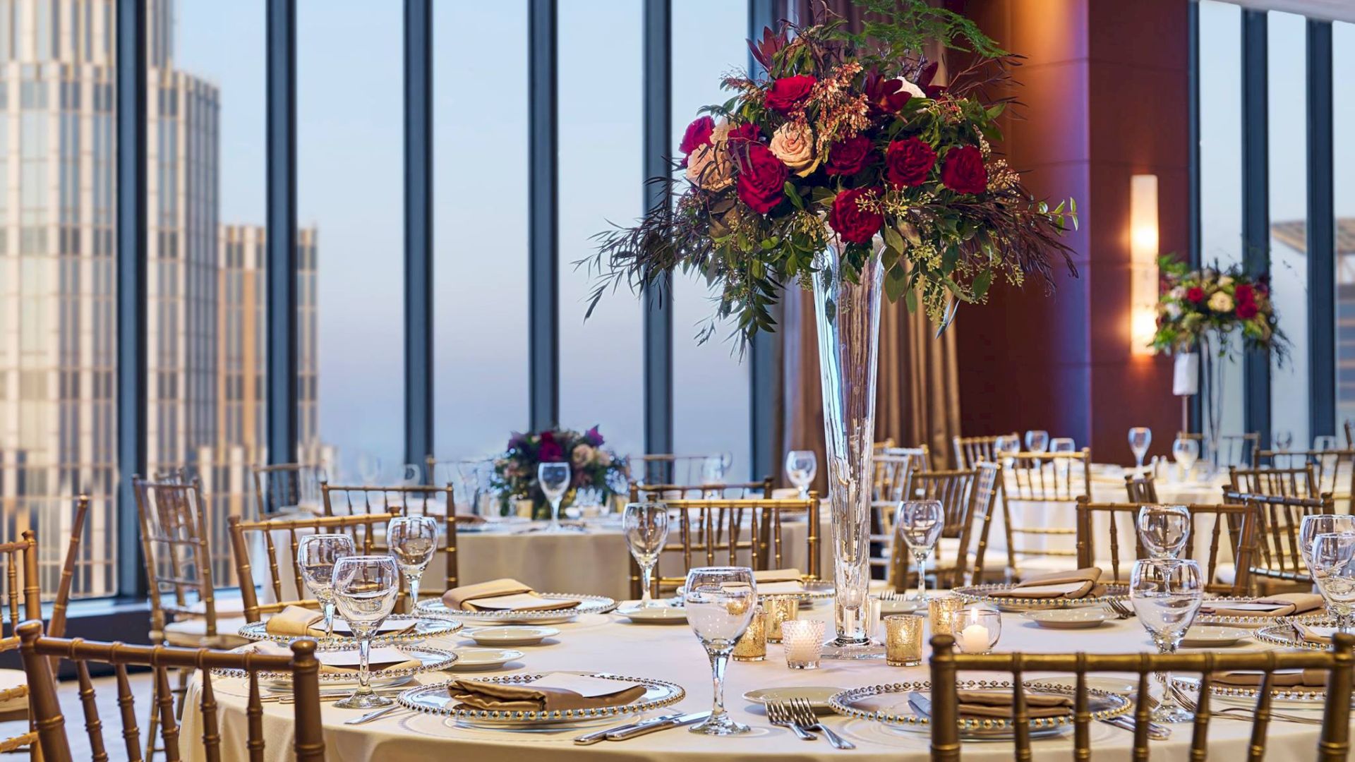 A lavish event table with gold chairs and candlelit place settings, topped by a large bouquet centerpiece of deep red flowers.