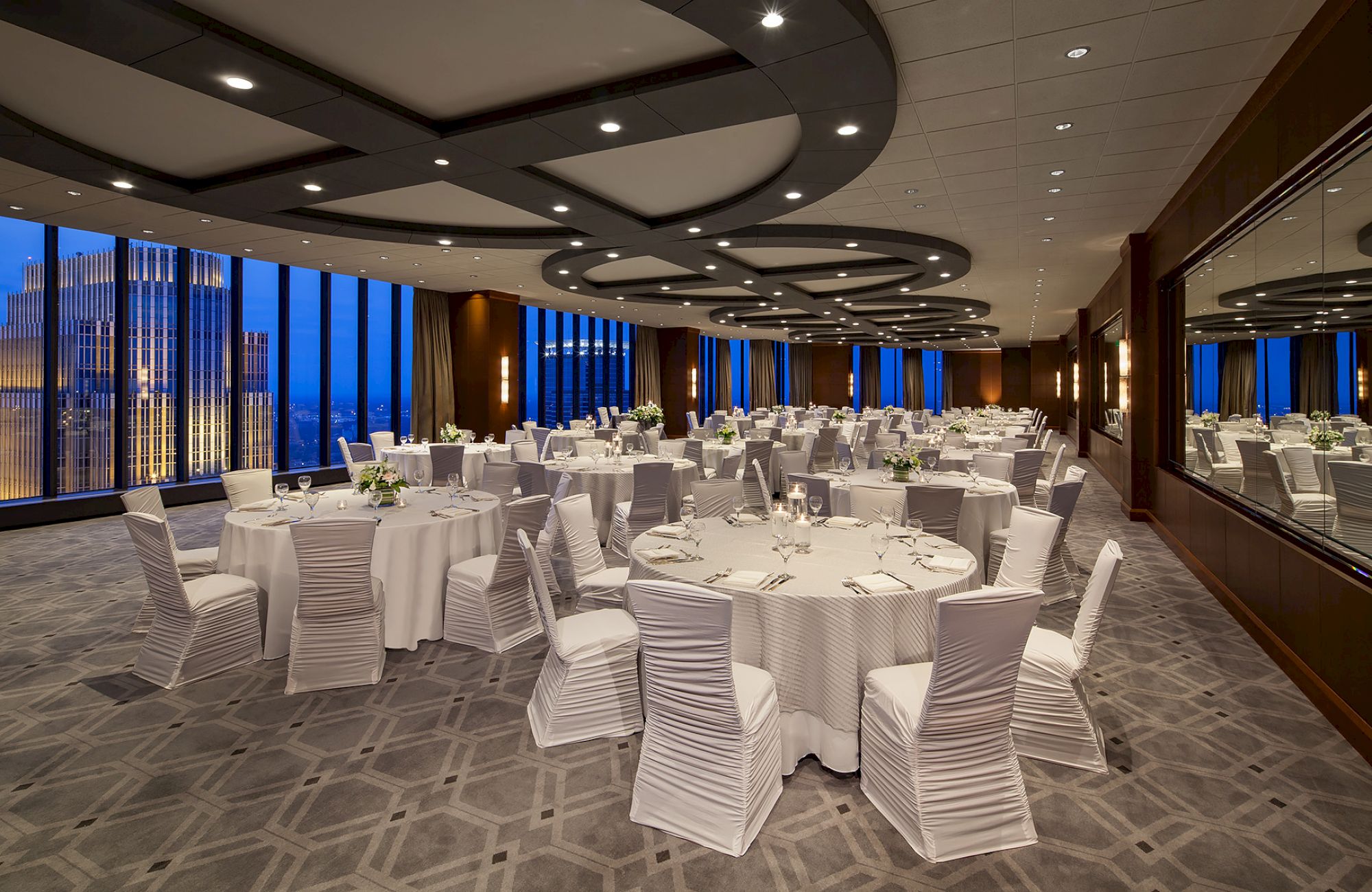 A spacious banquet hall set for a formal event, with round tables, white chair covers, soft lighting, and city lights visible through large windows.