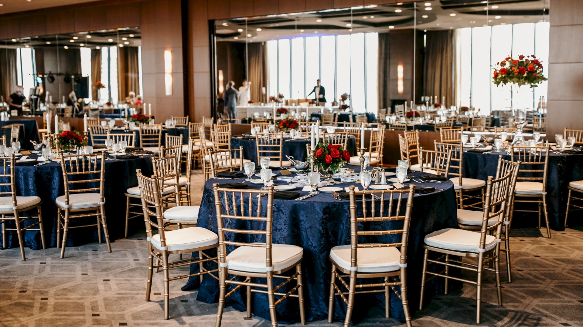 A banquet hall is set with round tables, gold chairs, place settings, candles, and red flower centerpieces for an event.